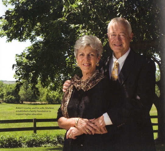 An elderly couple stands outside, smiling, with trees in the background. The woman has short gray hair and wears a scarf, while the man in a suit stands behind her, holding her shoulders.