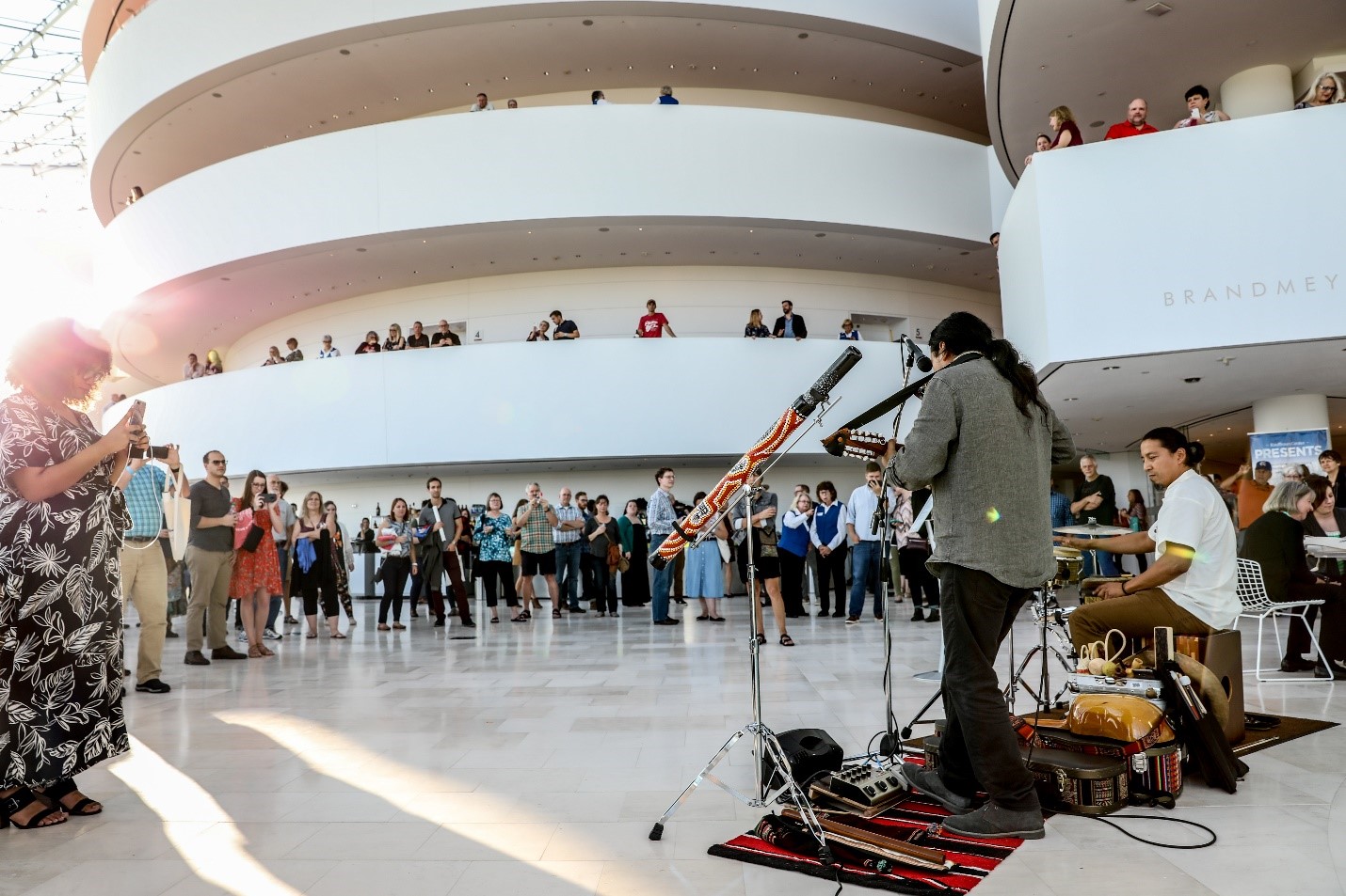 A musical duo performs on various instruments in an open indoor space as a crowd watches, with some people on multiple floors looking over railings.