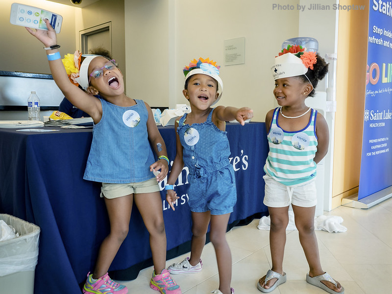 Three young girls stand together smiling and posing in front of a table with a blue tablecloth. They are wearing fun hats, casual clothes, and wristbands, with name tags on their outfits.