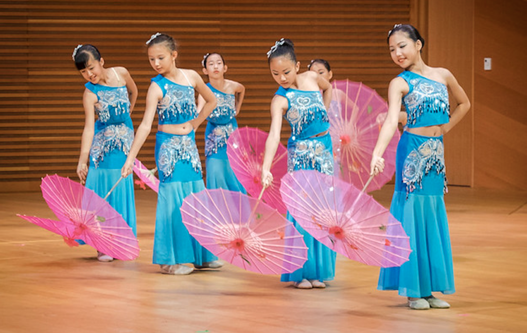 A group of young dancers in blue costumes perform with pink umbrellas on a wooden stage, showcasing Our Impact for the Performing Arts.