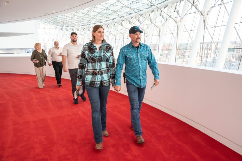 A group of people walk down a curved hallway with a red carpet. The hallway features large windows and modern architecture.