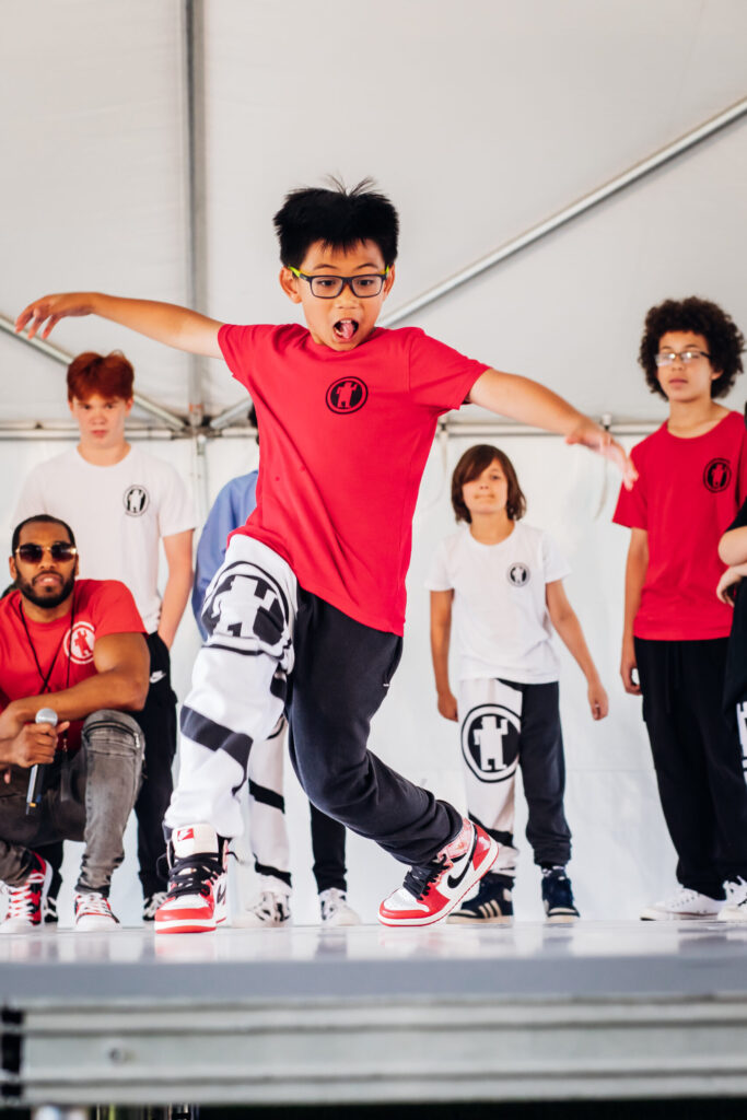 A boy wearing glasses and a red t-shirt excitedly performs a dance move on stage while others wearing white and red t-shirts look on. A seated man with a microphone is also visible in the background.