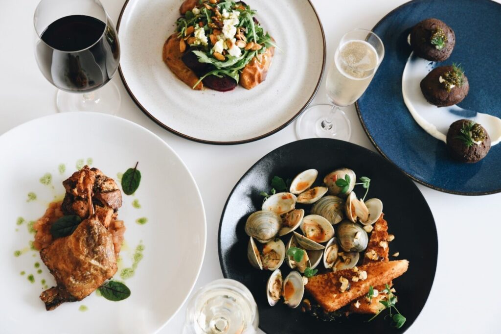 A table set with a variety of dishes, including a plate of clams, a roasted meat dish, a salad, two dark round desserts, and two glasses of beverages, one dark and one light.