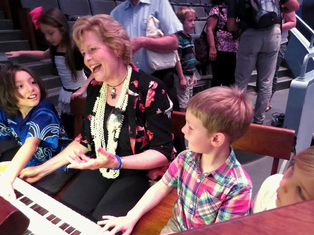 An older woman sits at a keyboard, smiling and interacting with excited children in a classroom or event setting. Other children and adults are in the background.