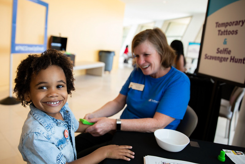 A child with curly hair smiles at the camera while seated at a table. An adult in a blue shirt is applying a temporary tattoo to the child's hand. Signs nearby mention temporary tattoos, a selfie scavenger hunt, and volunteer opportunities in Kansas City.