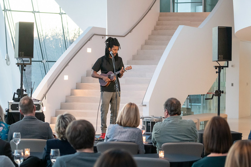 A person playing the guitar and singing on a small stage in front of an audience at an indoor event hosted by a Performing Arts Nonprofit in Kansas City, all amidst modern architecture.