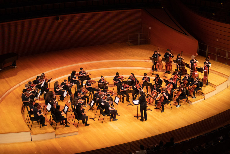 A symphony orchestra, including string sections and a conductor, performs on the wooden stage of the Kansas City Performing Arts concert hall, with tiered seating in the background.