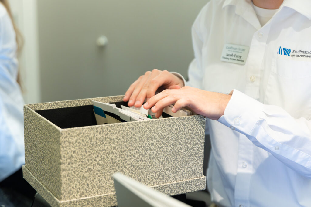 A person in a white shirt is sorting through a file box containing various folders and documents, including A Guide to the Kauffman Center.