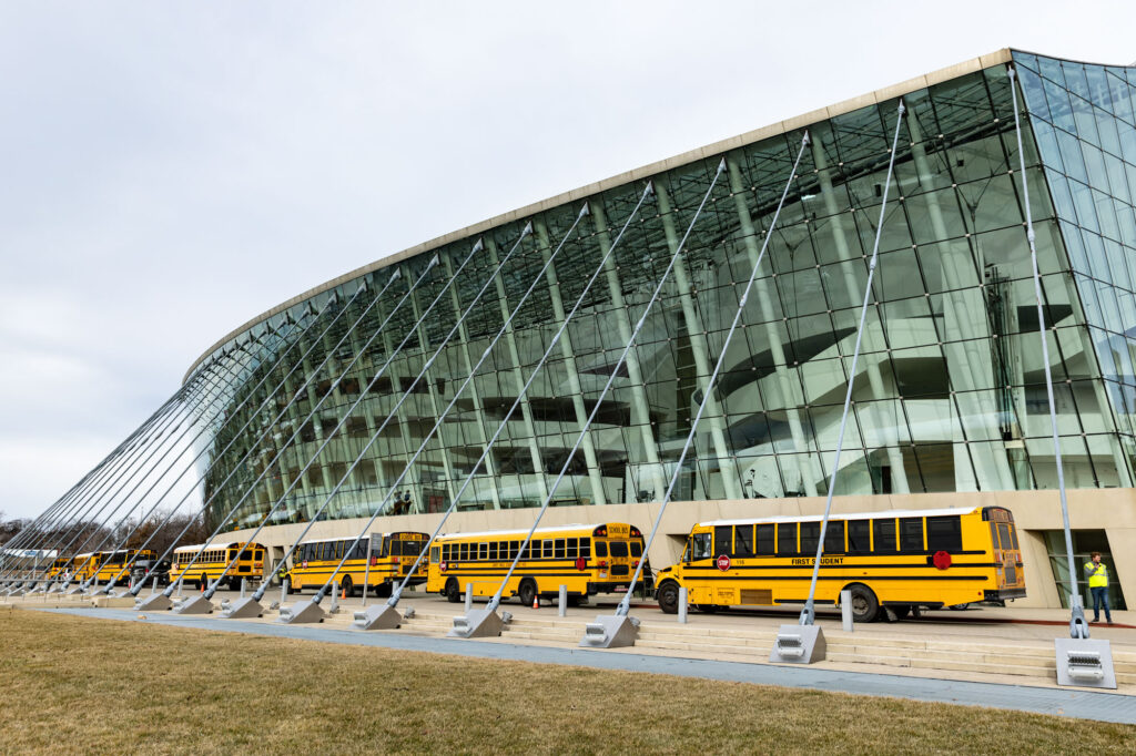 Several yellow school buses are parked in front of a modern glass building with a curved facade, as seen in at the Kauffman Center.