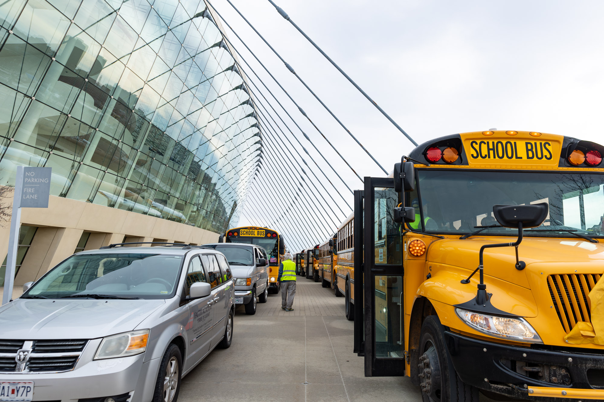 Parked school buses and cars line up in front of a modern glass building, while a person wearing a safety vest stands nearby.