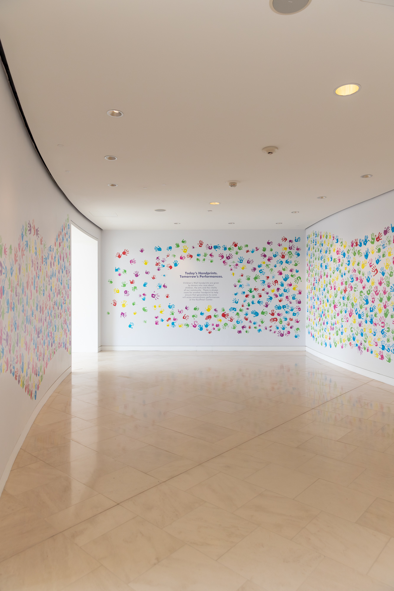 A wall featuring colorful handprints surrounds text explaining that children's handprints are donations supporting community arts, beautifully displayed at the Kauffman Center for the Performing Arts. Visitors can view this heartwarming exhibit as part of a guide to the Kauffman Center.