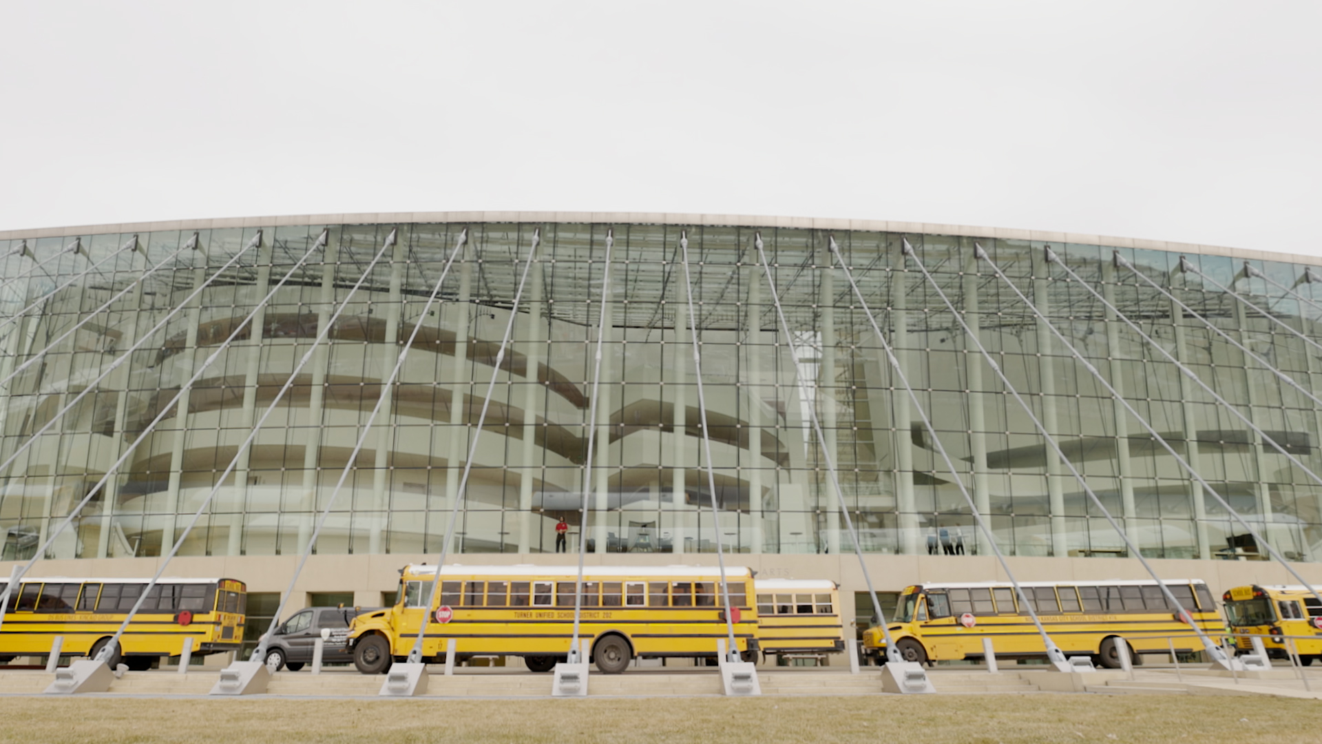 A modern glass building with a curved facade, likely the Kansas City Performing Arts center, is seen in the background. In front, three yellow school buses are parked in a row.