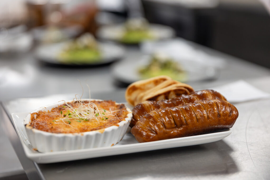 A white plate with a sausage, a side of pita bread, and a baked dish in a ceramic ramekin, garnished with sprouts—like an artsy dish