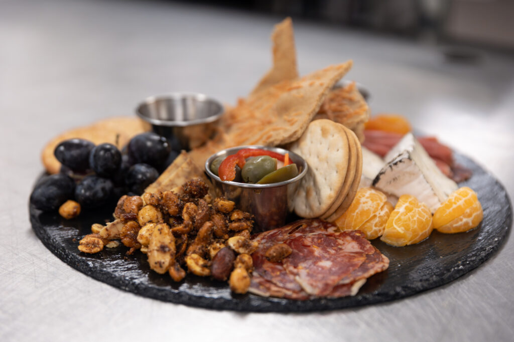 A charcuterie board with sliced meats, various cheeses, crackers, flatbreads, nuts, grapes, mandarin slices, and two metal condiment cups containing pickles and nuts