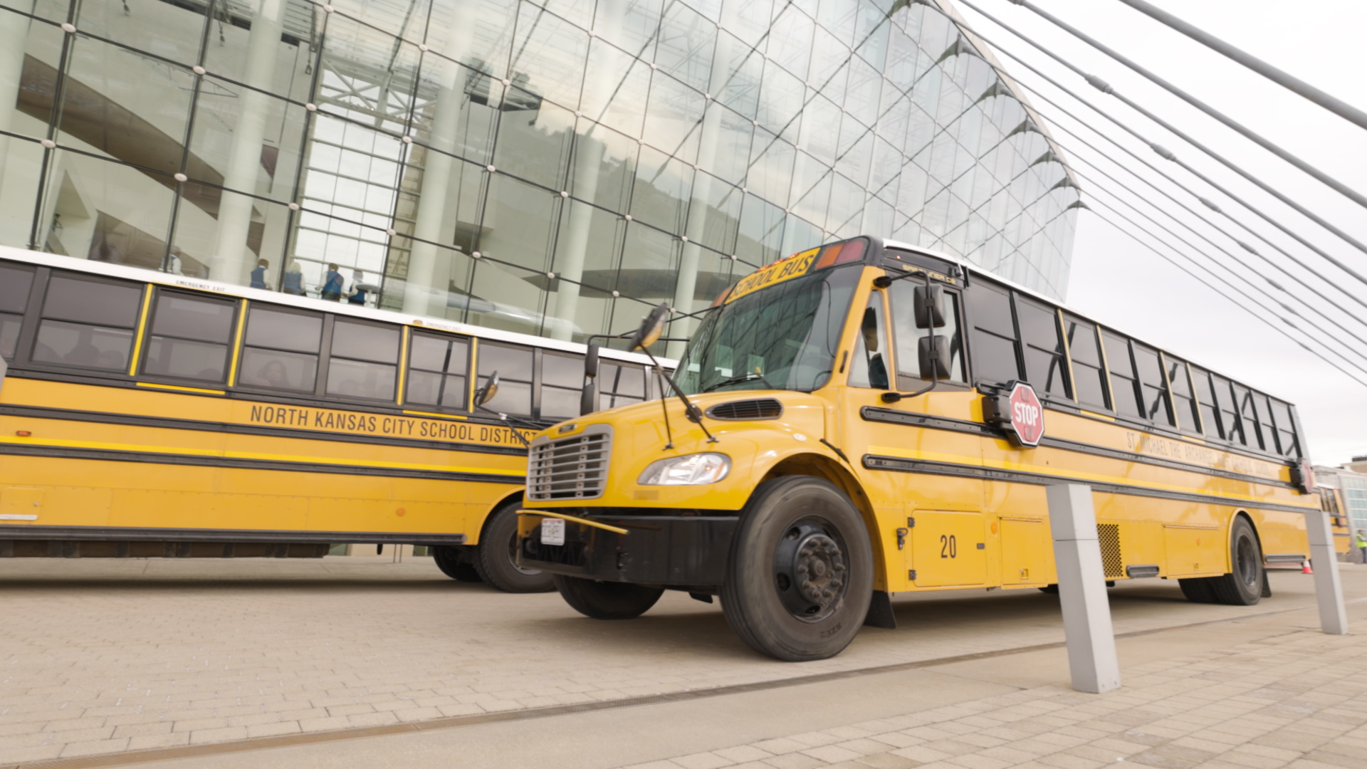 Two yellow school buses parked outside a modern glass building, marked with "North Kansas City School District" and featuring extended stop signs. The striking glass facade hints at the vibrancy within, perhaps a nod to the nearby Kansas City Performing Arts scene.