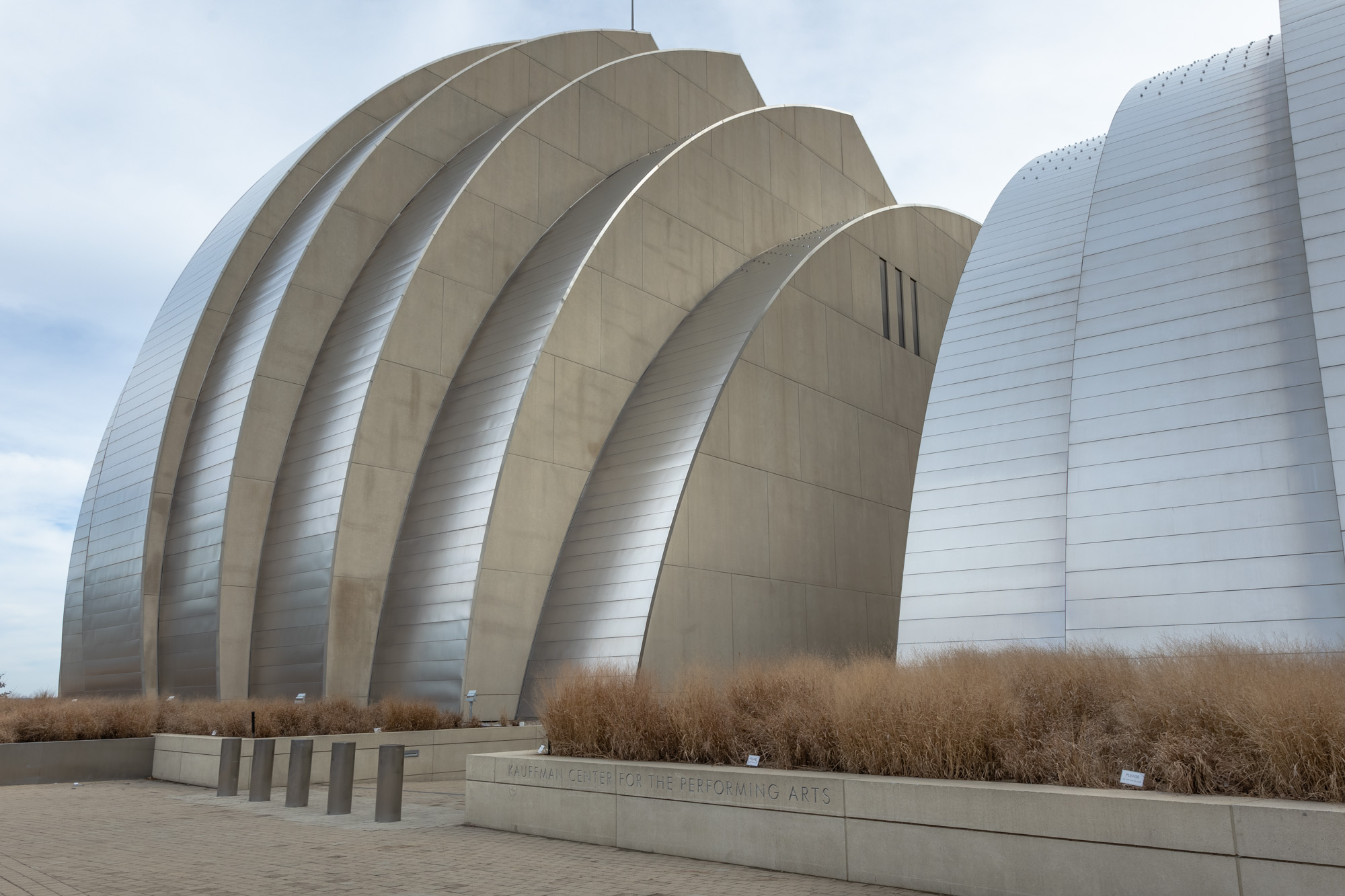 Modern architectural structure with a metallic, ribbed exterior and an inscription that reads "The Kauffman Center for the Performing Arts." Pathway and ornamental grass in the foreground.