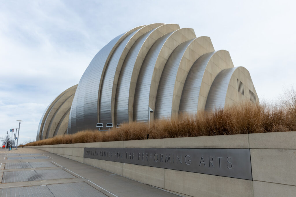 Modern architectural structure with a metallic, ribbed exterior and an inscription that reads "The Kauffman Center for the Performing Arts." Pathway and ornamental grass in the foreground.