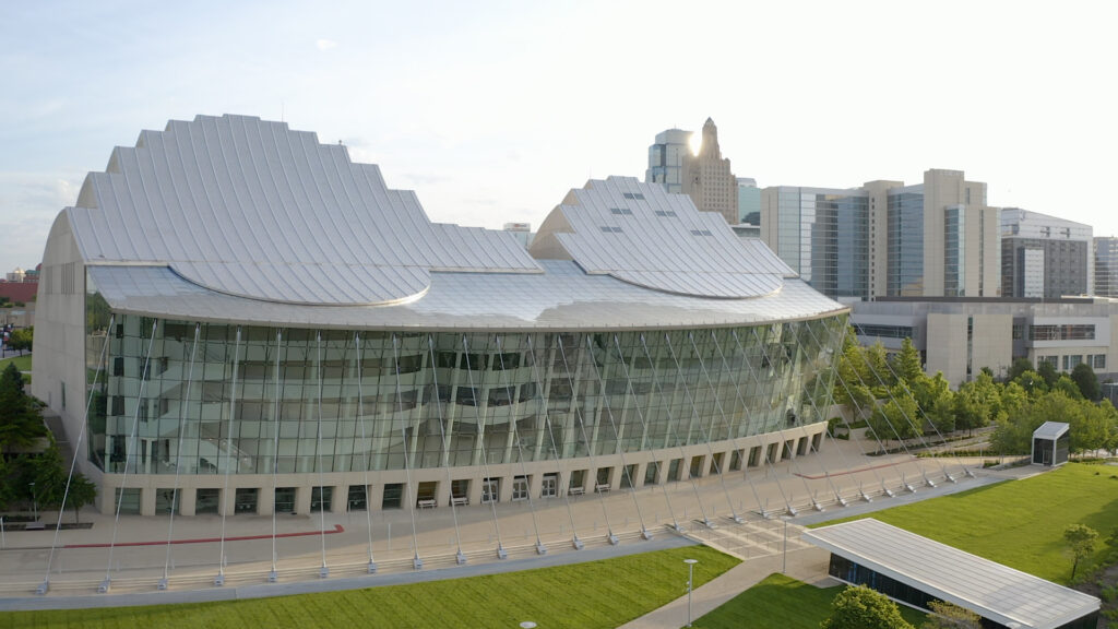 Modern building with a wavy glass façade and white roof, surrounded by a green lawn and city buildings in the background, the Kansas City Performing Arts Center.