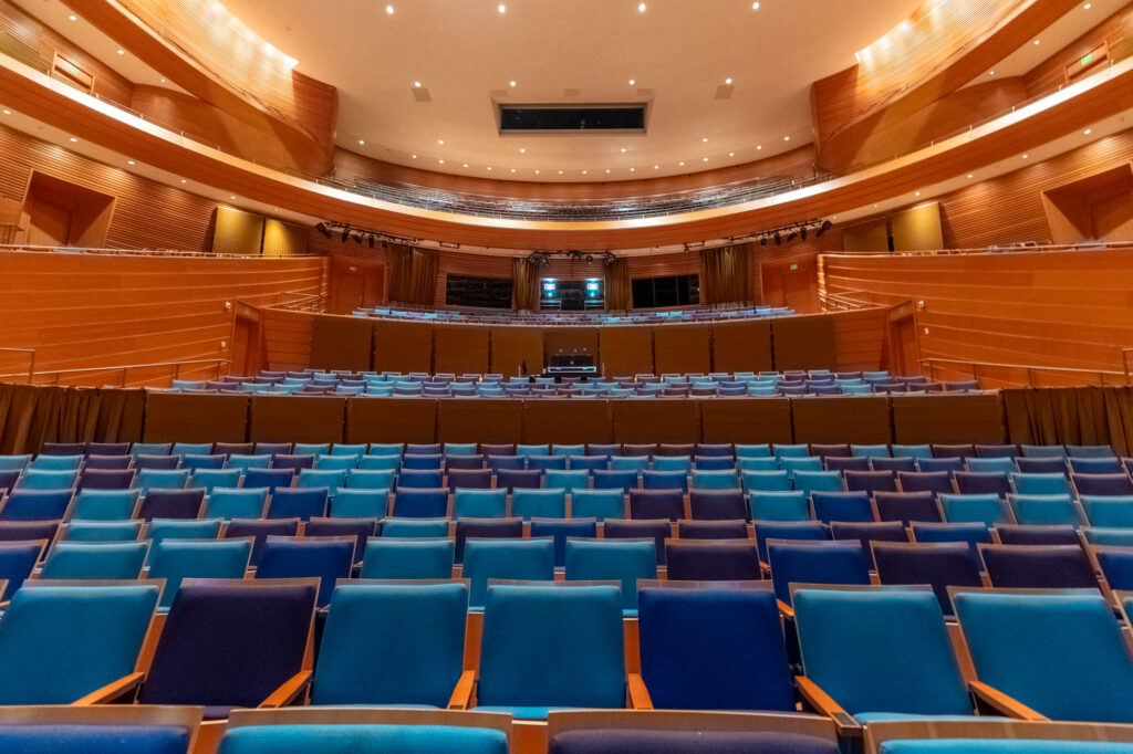 A modern, empty theater with blue and turquoise seats, wooden paneling, and a stage at the front under bright overhead lights—an architectural marvel.