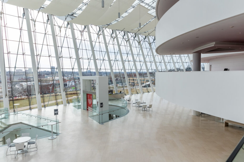 Spacious modern atrium with large windows, glass walls, and light beige flooring. Features a small seating area with white tables and chairs, and a white curved structure in the foreground.