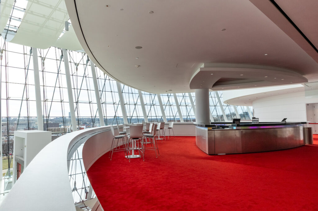 A modern interior space with large glass windows, a curved counter, and bar stools on a red carpet floor offers an inviting atmosphere.