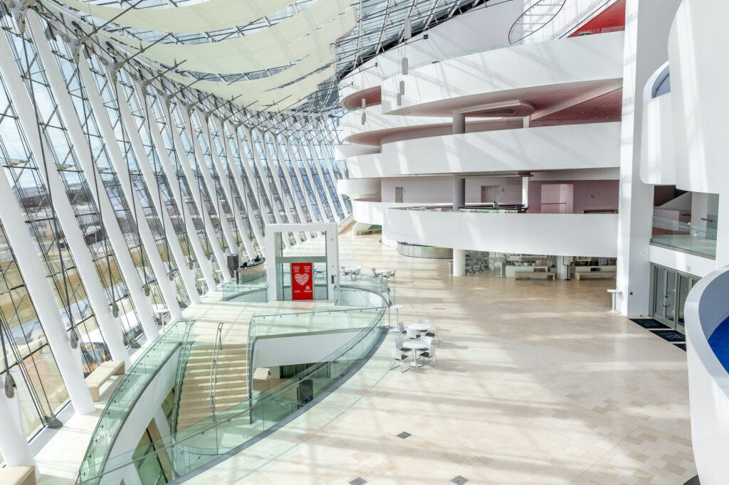 Modern, open-concept atrium with large glass walls, curved white balconies, a spiral staircase, and minimalist furnishings. Natural light floods the space, creating a bright and airy ambiance.