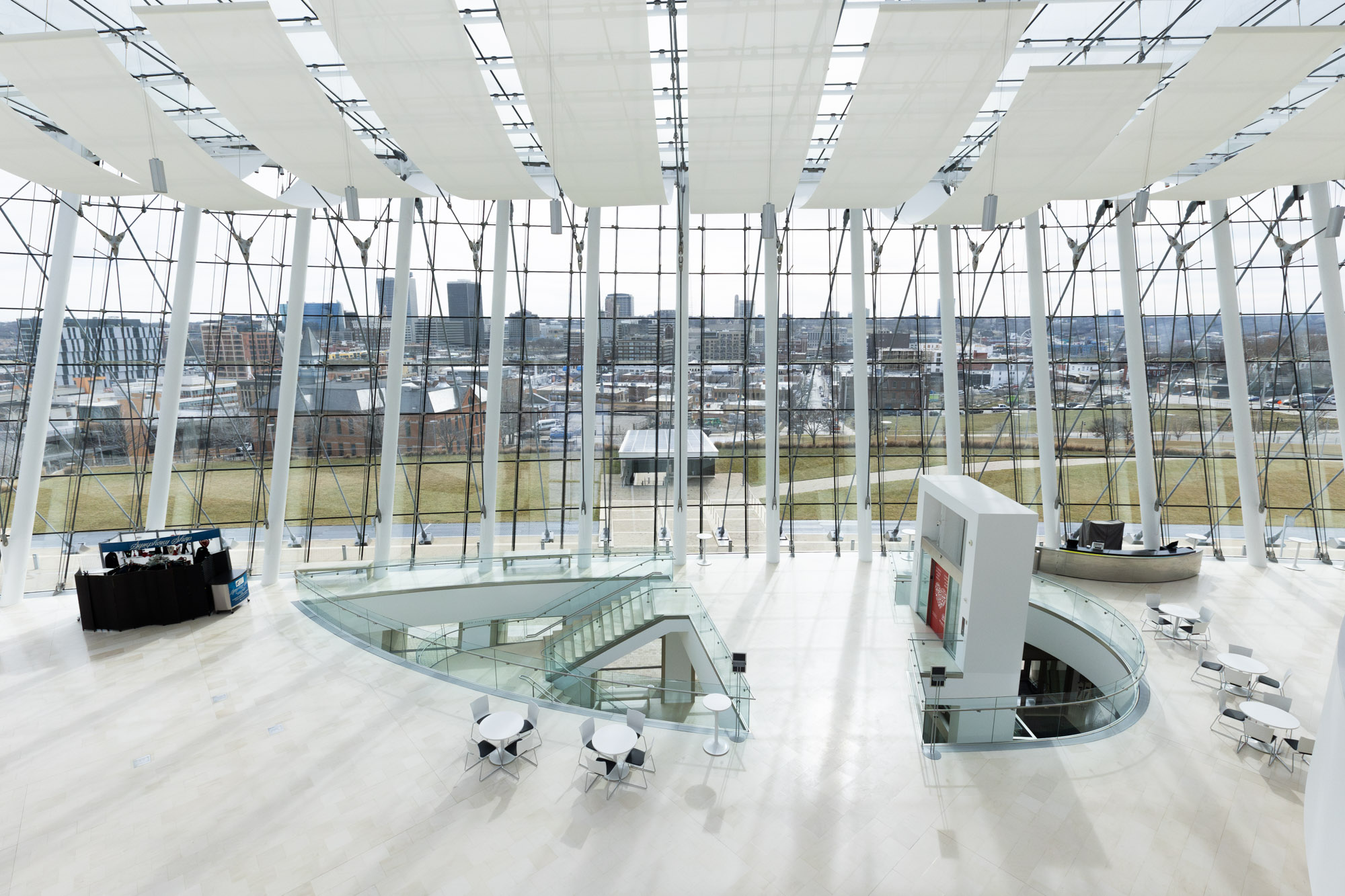 Spacious modern atrium with large windows, a few tables and chairs, and an open view of a cityscape in the background. The area features minimalist decor and ample natural light