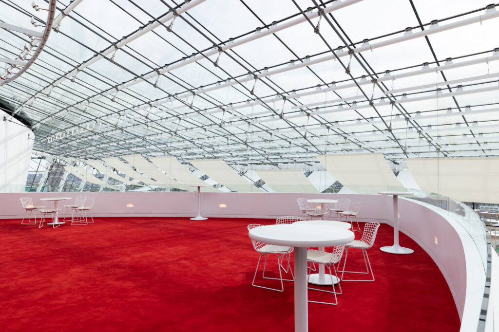 A modern indoor space with a glass ceiling, red carpet, and white tables and chairs, the area appears bright and airy.