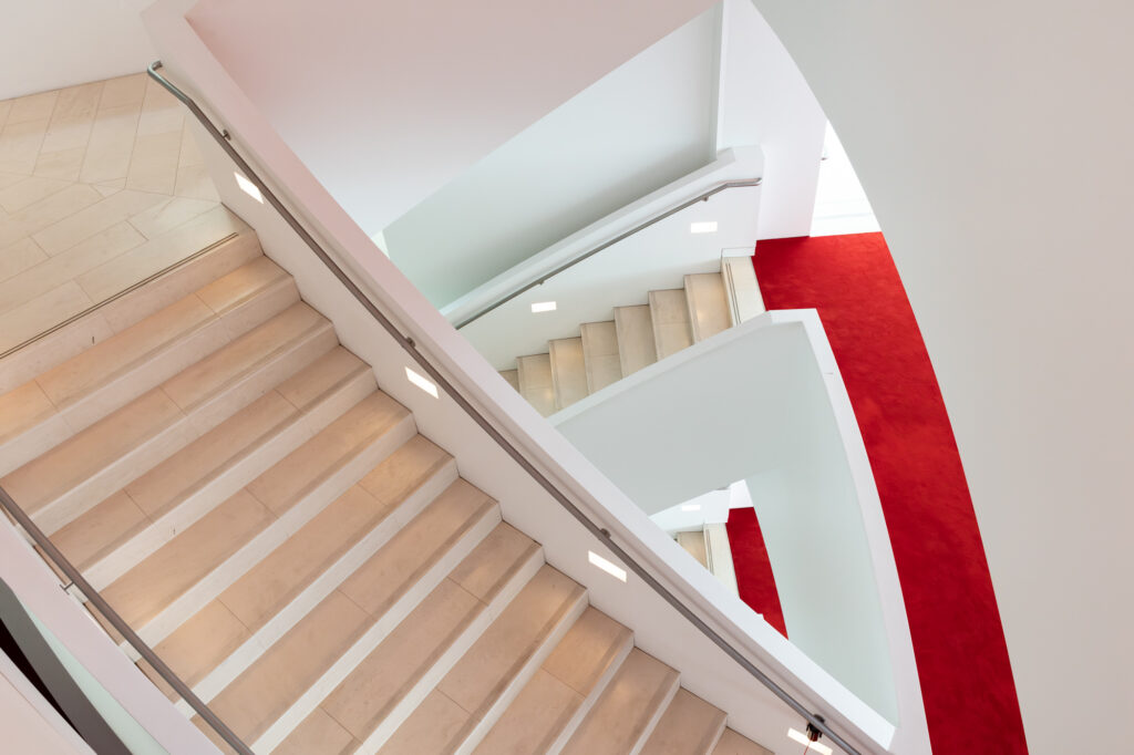 A spiral staircase with beige steps and white railings descends alongside a bright red carpet, illuminated by small wall-mounted lights