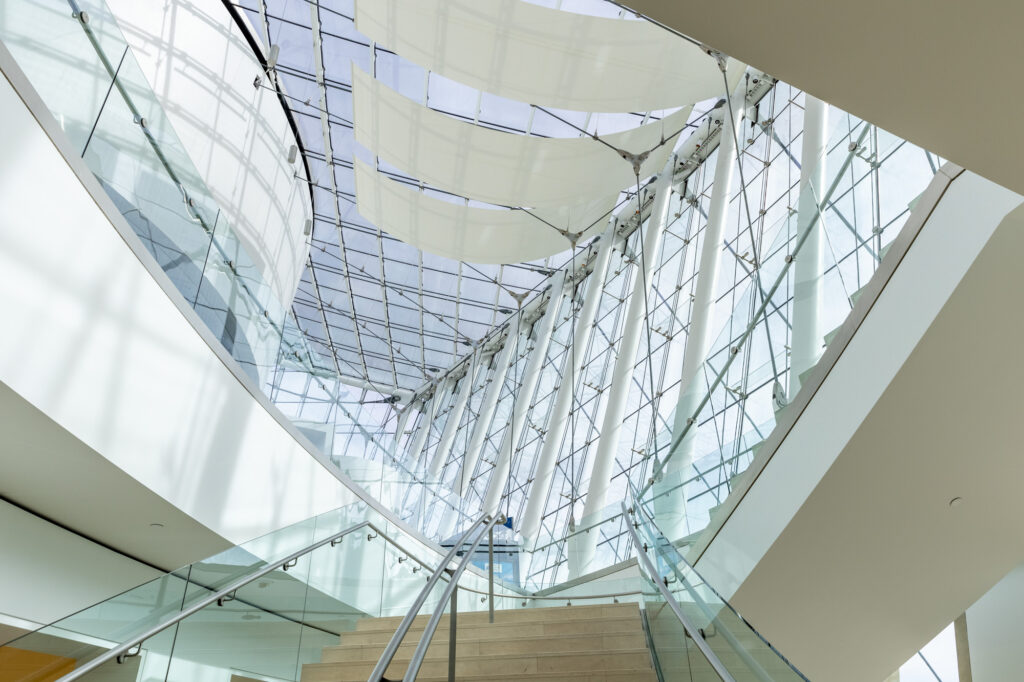 Modern architectural interior featuring a curved staircase with glass railings, sleek white walls, and an expansive glass ceiling with white fabric panels