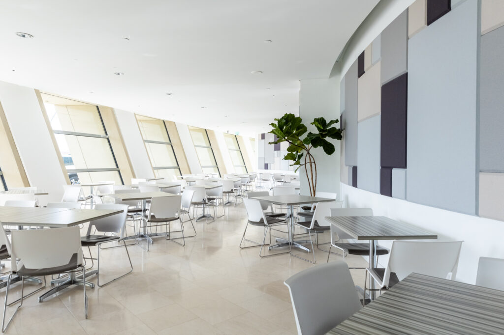 A modern cafeteria with white walls, large windows, and scattered tables with chairs. A tall plant is placed near a wall with geometric patterns. The space is clean and well-lit, much like the architectural elegance of the rest of the Kauffman Center.