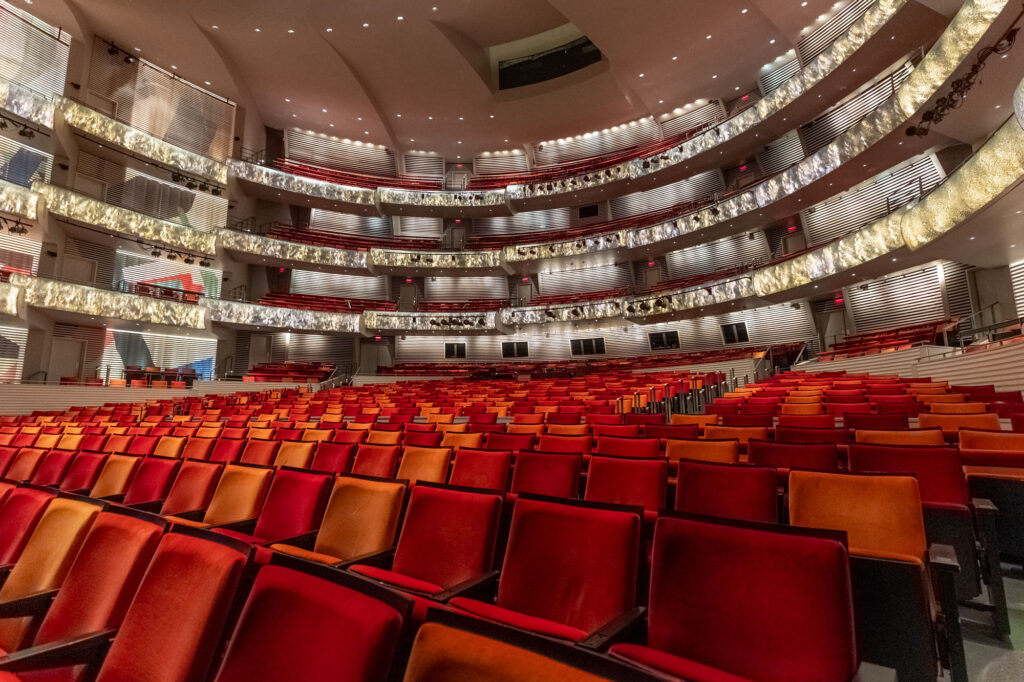 An empty theater with rows of red and orange seats, multiple balconies, and modern architectural design of the Kauffman Center.