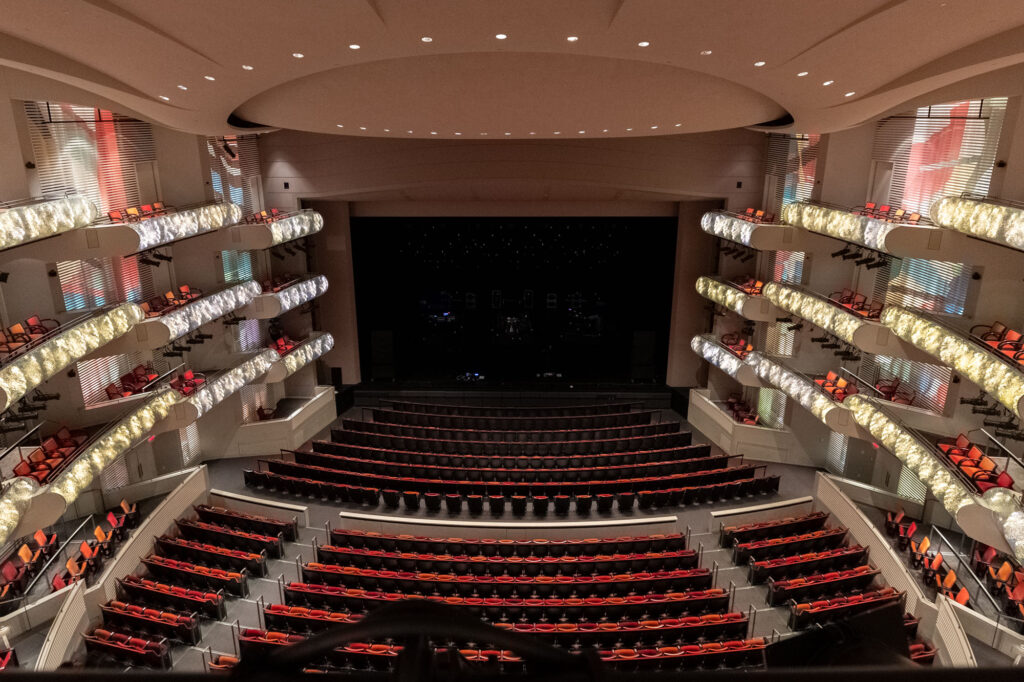 A view of an empty theater auditorium with multiple rows of red, cushioned seats and a modern stage setup, taken from the upper balcony at the Kauffman Center.