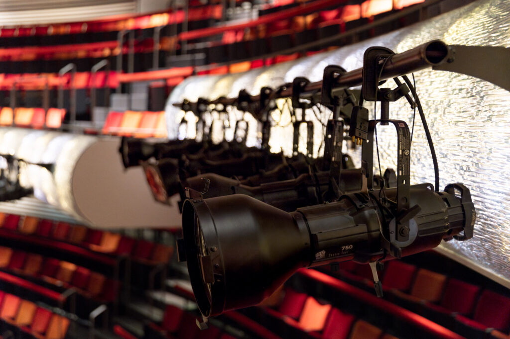 Stage lights hang from a fixture, focused on the seating area of a theater with rows of red and orange seats, reminiscent of a grand performance highlighted at the Kauffman Center.