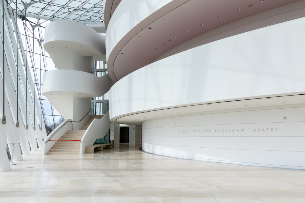 Spiral staircase and sleek modern architecture define this interior space. A sign on the wall reads "Muriel McBrien Kauffman Theatre." Large windows and a glass ceiling allow natural light to flood the area, offering a glimpse into the Kauffman Center's architectural brilliance.