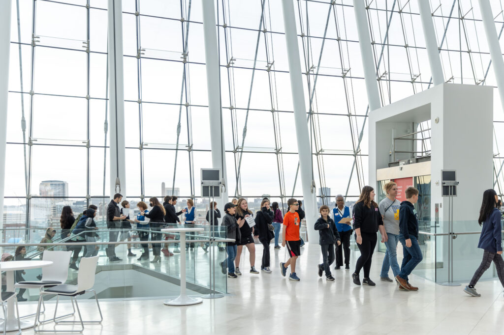 People walking and standing inside a modern, glass-walled building with a city view in the background, inside the Kauffman Center.