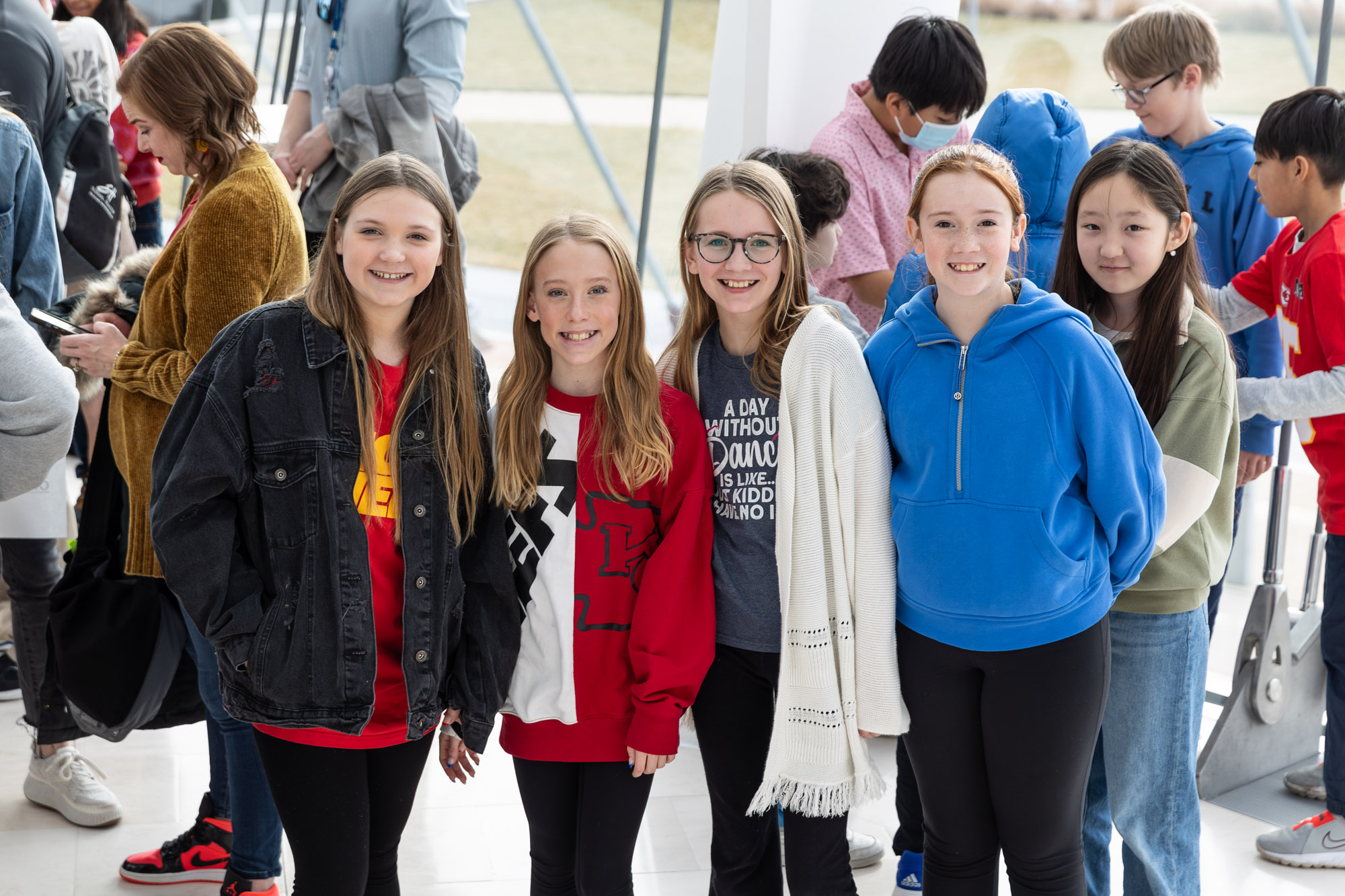 A group of six young individuals stands indoors, smiling at the camera. Four of them are in the foreground, dressed casually, while two more are in the background, engaged in conversation. at the Kauffman Center.