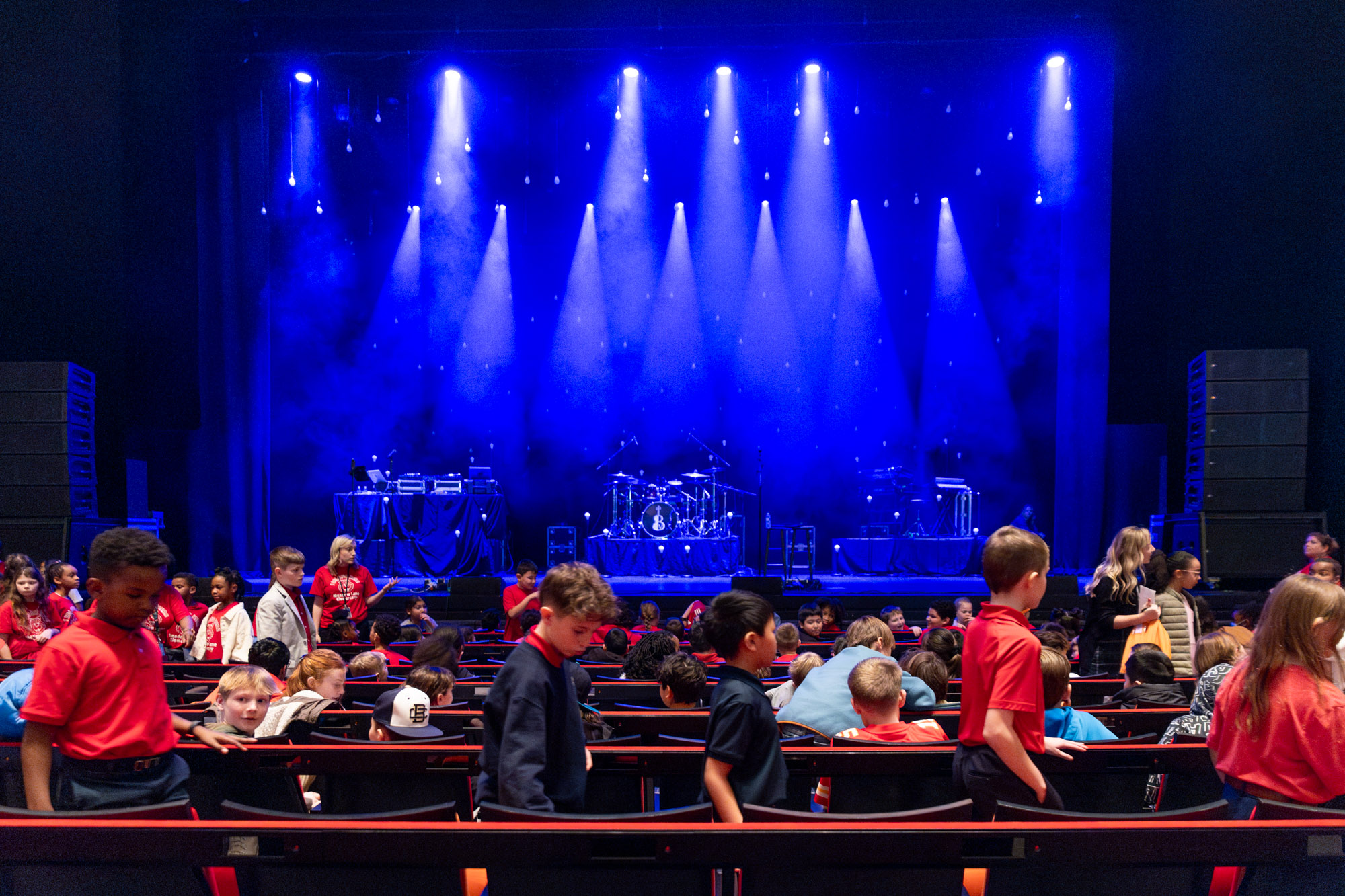 Children gather in an auditorium with a blue-lit stage featuring musical instruments and equipment for an upcoming performance, reminiscent of scenes from "A Guide to the Kauffman Center.