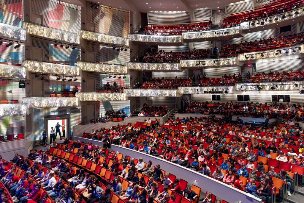 A large auditorium with multiple levels of seating is filled with an audience. The venue features modern design elements and is well-lit, embodying the elegance of the Kauffman Center.