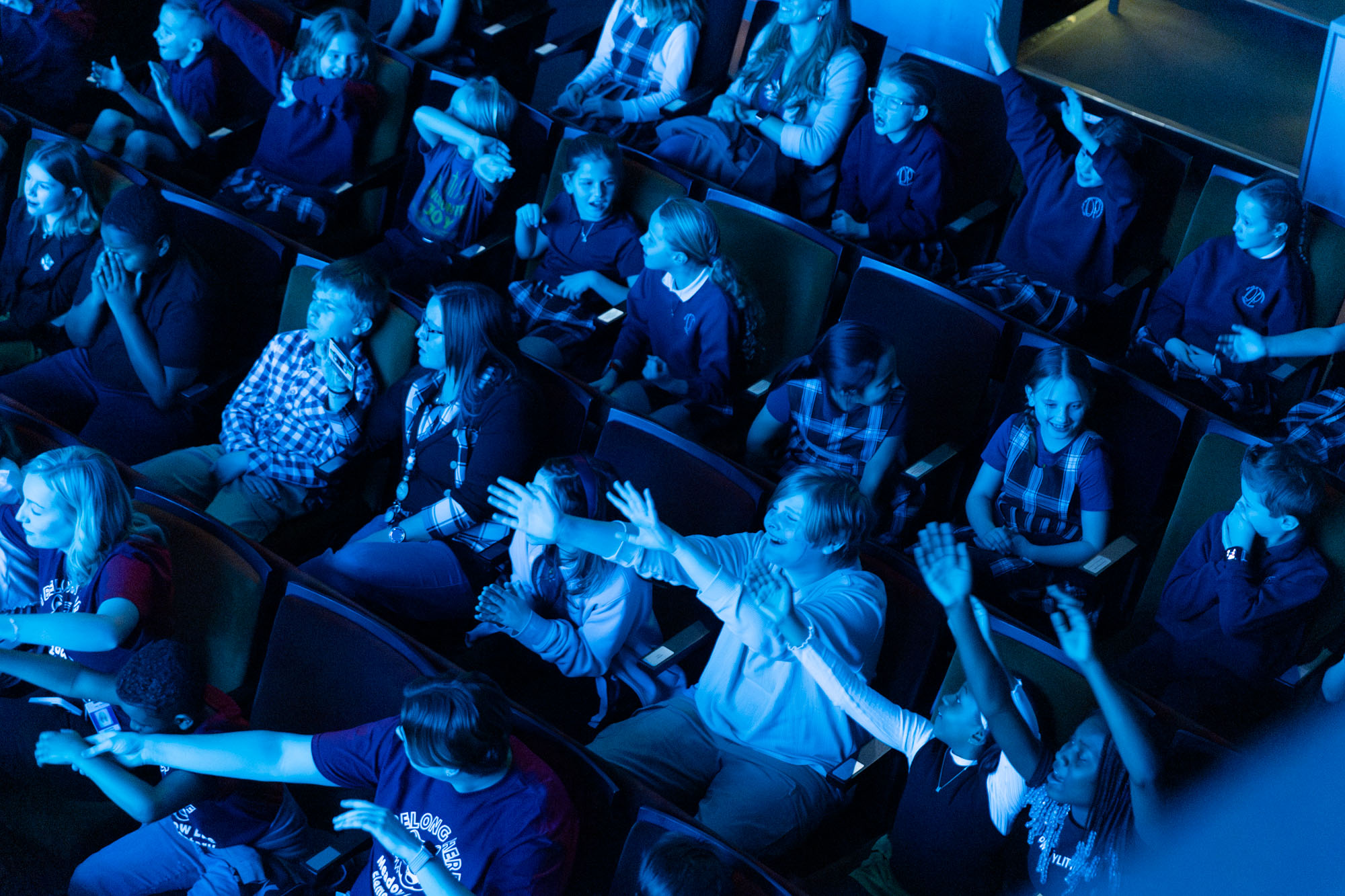 a group of children and adults sit in theater seats bathed in blue lighting. Some appear excited, raising their arms towards the stage, while others sit calmly.