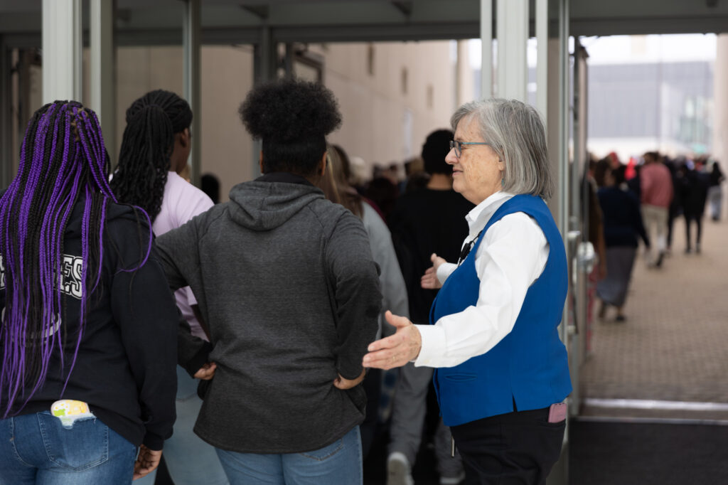 An older woman in a blue vest, appearing as a guide to the Kauffman Center, directs a group of people through glass double doors in a busy indoor setting.