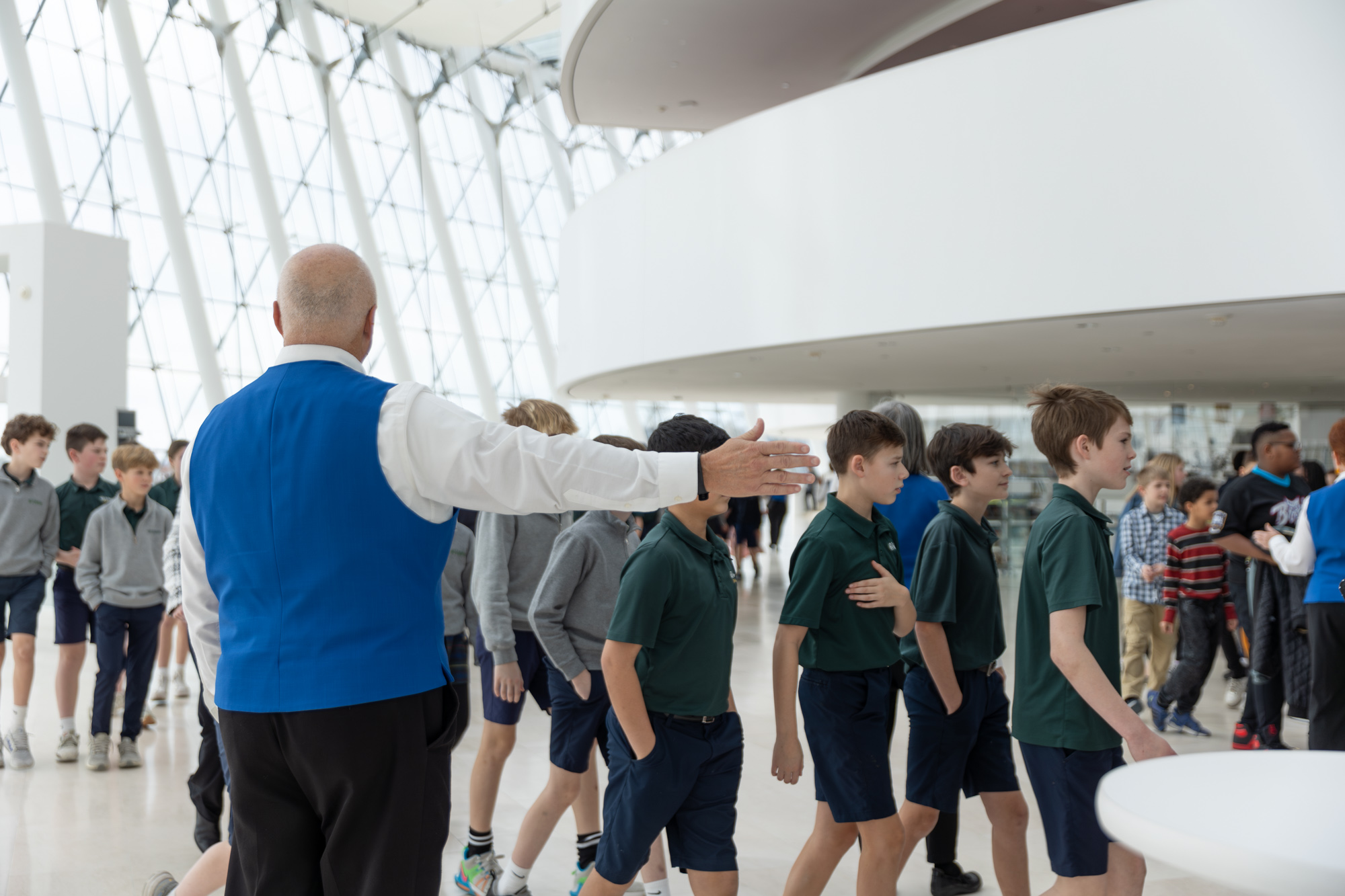 A man in a blue vest gestures toward a group of students in uniforms walking inside the large, modern Kauffman Center with its curved ceiling and expansive windows.