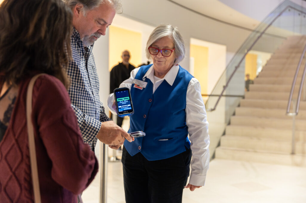 A ticketing attendant in a blue vest scans a man's ticket with a handheld device while another person watches at the entrance of the building, as if turning to "A Guide to the Kauffman Center" for navigating its grand staircase in the background.