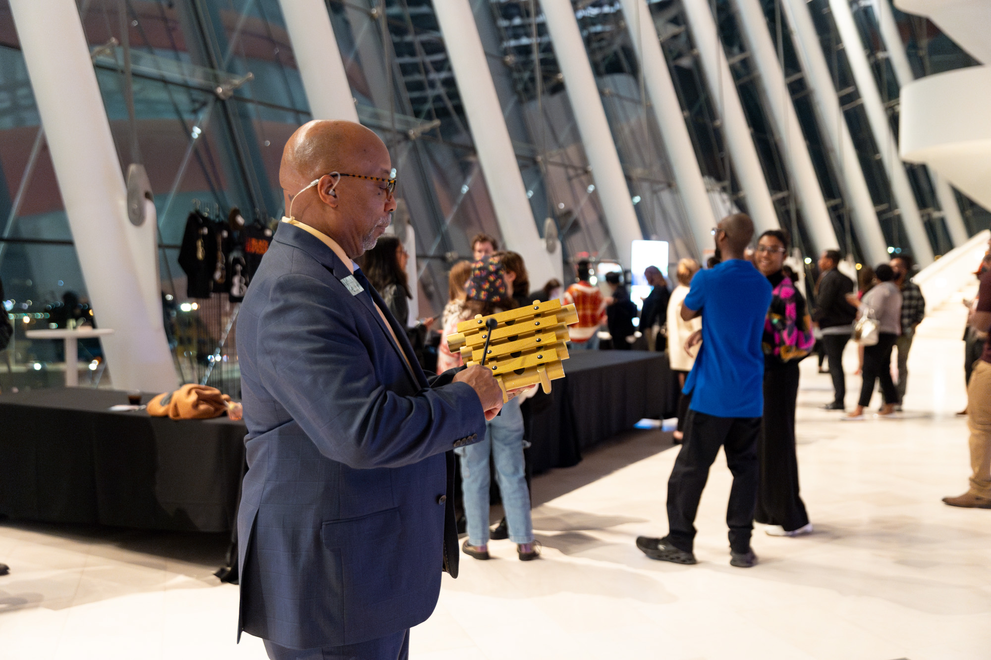 A man in a suit plays a xylophone in a large, modern indoor space at the Kauffman Center, with people standing and conversing in the background.