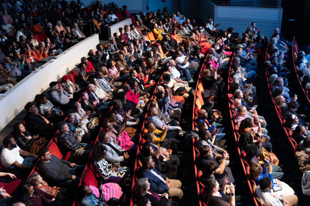 A diverse audience seated in a large theater, attentively facing the stage to the right. The theater seats are arranged in rows, and the patrons are dressed in a variety of clothing styles, creating a vibrant atmosphere described in A Guide to the Kauffman Center.