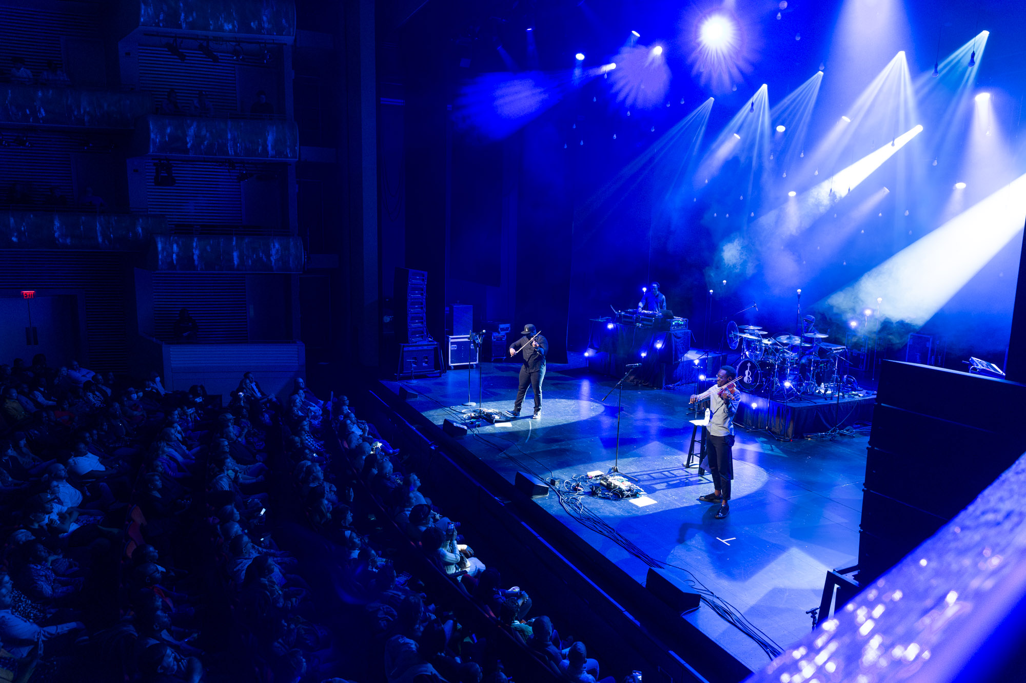 A band performs on a dimly lit stage with blue lighting in the theater, creating an atmosphere reminiscent of A Guide to the Kauffman Center, while an audience watches from tiered seating.