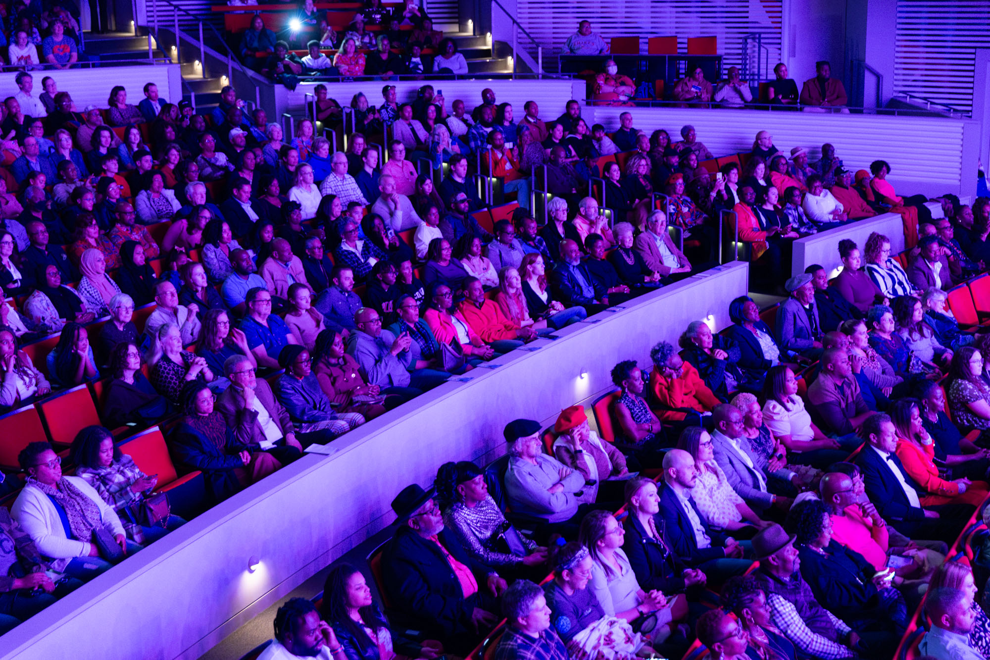 A large audience sits attentively in a tiered, modern theater, illuminated by purple lighting, reminiscent of what you might find in A Guide to the Kauffman Center.