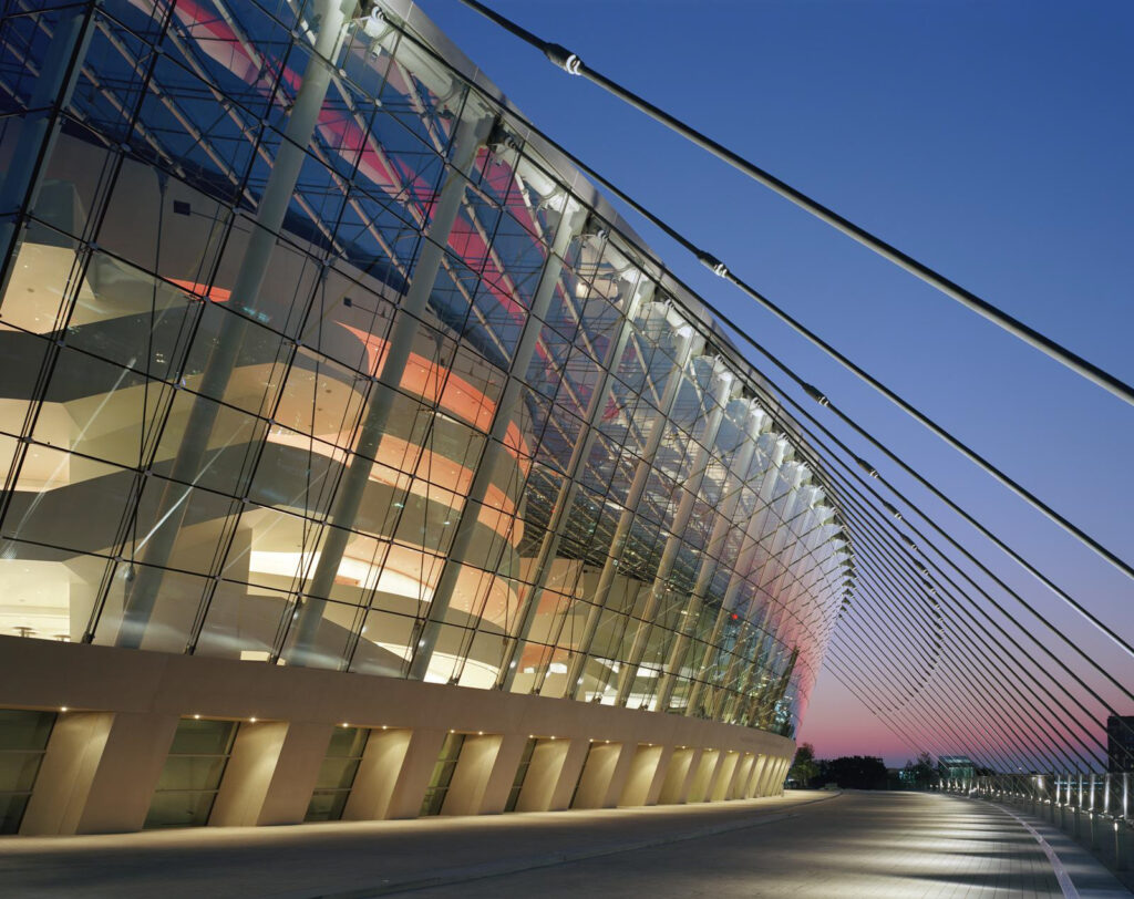 A modern glass building with a curved facade and exterior support cables is illuminated at dusk, evoking the sleek design of the Rent Kauffman Center.