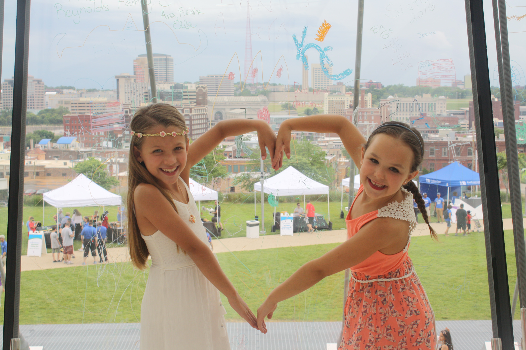 Two smiling young girls in summer dresses form a heart shape with their arms in front of a large window, overlooking the bustling Future Stages Festival with tents and the cityscape in the background.