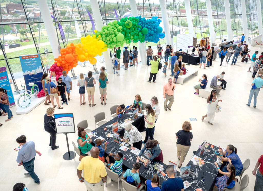 A large group of people participates in various activities inside a bright, spacious building. A colorful balloon arch and large windows are visible, with some people gathered at tables and others standing around, contributing to our Impact for the Performing Arts.